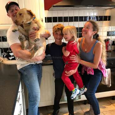 Das Bild zeigt eine vierköpfige Familie, darunter zwei Kinder und ein Hund, die für ein Foto in einer Küche mit weißen Schränken und schwarzen Arbeitsplatten posieren.