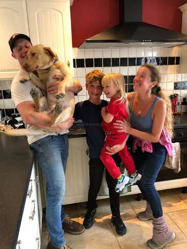 Das Bild zeigt eine vierköpfige Familie, darunter zwei Kinder und ein Hund, die für ein Foto in einer Küche mit weißen Schränken und schwarzen Arbeitsplatten posieren.