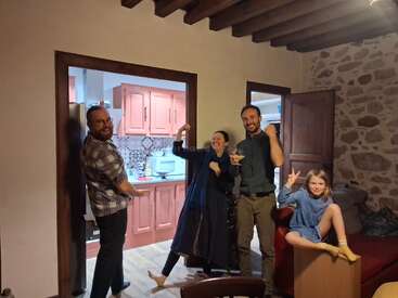 Four adults and a child pose playfully in a cozy living room with stone walls and wooden beams. The mood is joyful, relaxed, and celebratory.