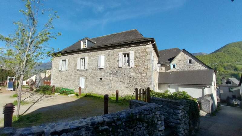 The image depicts a stone building with white windows and a dark roof, set against a blue sky with a mountainous backdrop.
