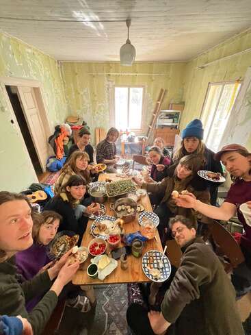 Um grupo de pessoas se reúne em torno de uma mesa de madeira, desfrutando de uma refeição em uma sala rústica com paredes amarelas desbotadas, compartilhando comida e conversando alegremente.