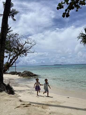 Deux jeunes enfants courent joyeusement sur une plage tropicale de sable fin, entourés d'une eau turquoise cristalline, de palmiers et d'un ciel bleu paisible et nuageux. Le bonheur à l'état pur.
