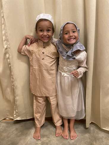 Deux jeunes enfants, un garçon et une fille, posent joyeusement côte à côte en costume traditionnel, un large sourire aux lèvres, les bras autour l'un de l'autre, devant des rideaux couleur crème.