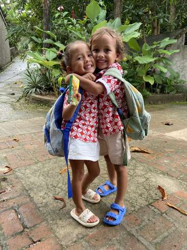 Deux jeunes enfants, vêtus de t-shirts à motifs rouges assortis et portant des sacs à dos de la même couleur, s'étreignent et sourient joyeusement en plein air, sur un sentier entouré de végétation luxuriante et d'arbres.