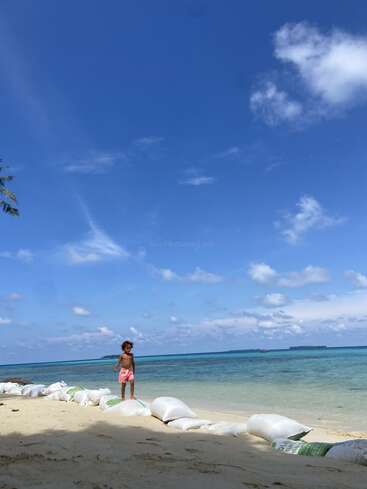 Un enfant se tient debout sur des sacs de sable, sur une magnifique plage tropicale, et contemple l'océan turquoise sous un vaste ciel bleu parsemé de nuages blancs.