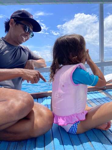 Un homme portant des lunettes de soleil et une casquette est assis en tailleur ; il sourit et montre du doigt une petite fille vêtue d'un gilet de sauvetage rose, lors d'une balade en bateau sous le soleil.