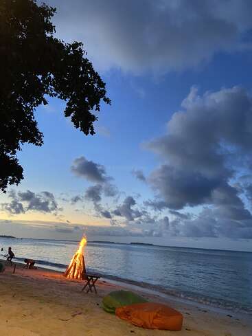 Une scène paisible sur la plage au coucher du soleil montre un feu de joie qui brûle près du rivage, des poufs et des tables posés sur le sable, sous un ciel spectaculaire parsemé de nuages.