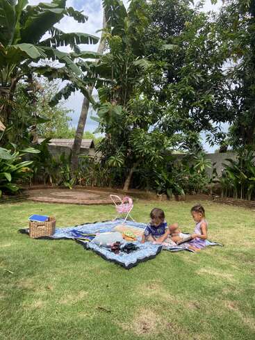 Deux jeunes enfants sont assis sur une couverture à motifs lors d'un pique-nique dans un jardin verdoyant et luxuriant, entouré de plantes tropicales, avec des jouets et un panier à proximité.