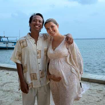 Un couple souriant se tient sur une plage de sable au bord de la mer, avec un bateau en arrière-plan. La femme semble enceinte ; tous deux ont l'air heureux et détendus.