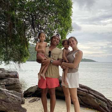 Uma família feliz de quatro pessoas está em uma praia arenosa perto de rochas e da água, cercada por uma vegetação exuberante, aproveitando juntos um dia tranquilo, tropical e nublado.