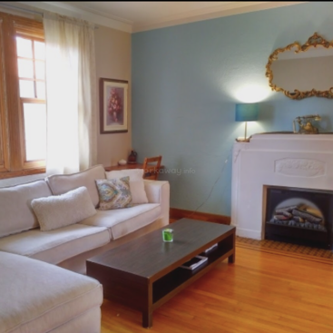 A cozy living room features a white sectional sofa, light blue accent wall, elegant fireplace, ornate gold mirror, wooden coffee table, and wooden floor. Warm atmosphere.