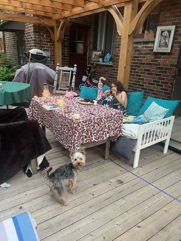 A cozy outdoor patio setup with a table covered in a patterned cloth, candles, two people relaxing on a bench, and a small dog looking up.