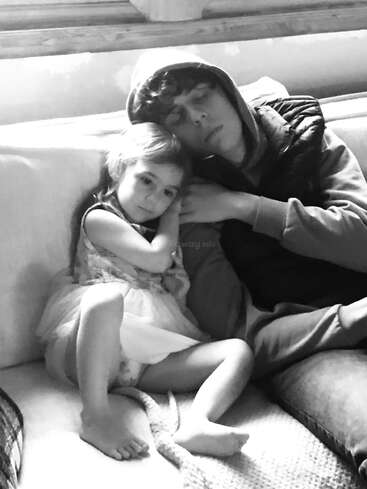 A young girl and an older boy, possibly siblings, cuddle together on a couch. The photo is black and white, conveying warmth, comfort, and familial love.