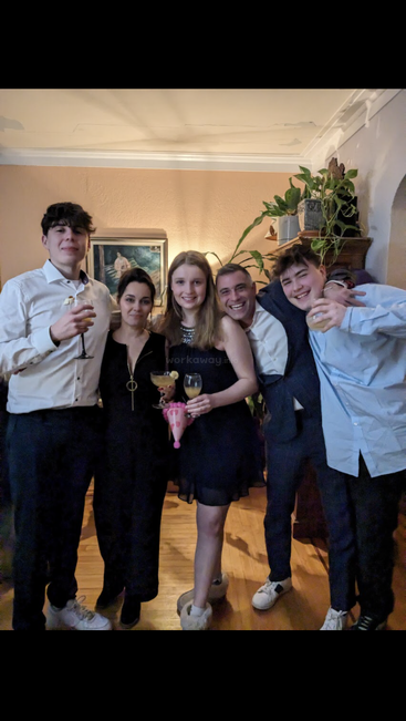 Five young people, dressed semi-formally, stand together smiling and holding drinks at a house party. There are green plants and a painting in the background.