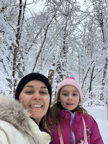 A woman and a young girl smile in a snowy forest. Both wear winter jackets and hats, surrounded by snow-covered trees and enjoying a cold, beautiful day.
