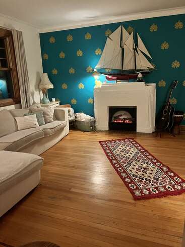 A cozy living room with a white sectional sofa, teal accent wall, fireplace, large sailboat model, patterned rug, guitar, and warm wooden floors.