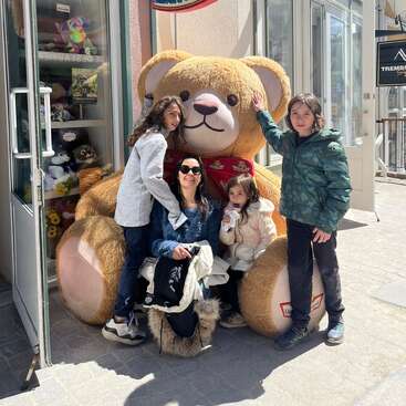 Una mujer y tres niños posan felices delante de una tienda junto a un osito de peluche gigante. Los niños sonríen y juegan entre ellos, creando un ambiente alegre y entrañable.