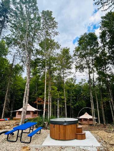 Un tranquilo camping en el bosque cuenta con dos tiendas de lona, una mesa de picnic azul brillante y una bañera de hidromasaje redonda de madera rodeada de altos árboles bajo un cielo parcialmente nublado.