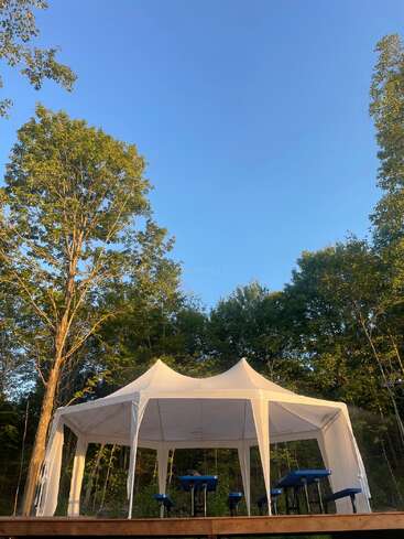 Una carpa blanca con toldo se erige sobre una plataforma de madera en un claro del bosque, rodeada de árboles verdes y bajo un cielo azul claro, con bancos debajo.