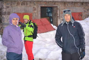 The image shows three individuals standing in front of a building, with two of them wearing matching outfits and the third wearing a different outfit, all facing the camera.