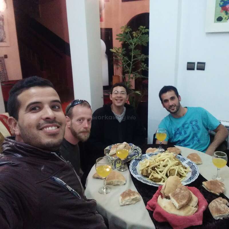 The image shows four men sitting at a table with food and drinks, posing for a selfie in what appears to be a restaurant or home dining area.