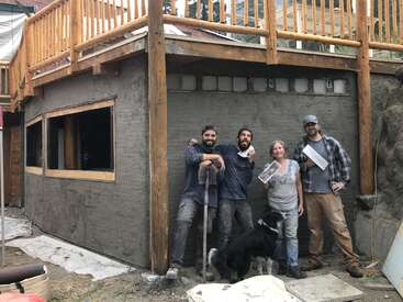 A imagem mostra quatro pessoas e um cachorro em frente a uma casa em construção, com o prédio apresentando vigas de madeira e um deck.