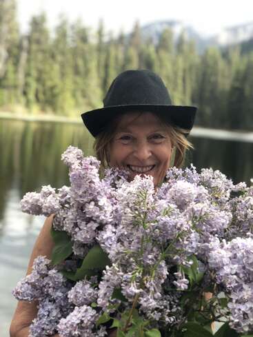 Uma mulher sorridente, com um chapéu preto, está à beira de um lago tranquilo, segurando alegremente um grande buquê de flores lilás, com árvores e montanhas ao fundo.