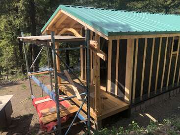 Uma pequena cabana de madeira em construção fica no meio da floresta. Há andaimes na entrada, e o telhado é feito de chapas de metal verdes.