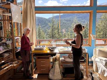Duas mulheres estão em uma sala aconchegante e bem iluminada, com janelas amplas. Elas conversam e sorriem, cercadas por plantas, estantes e uma vista deslumbrante da floresta e das montanhas.