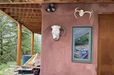 Uma varanda rústica com vigas de madeira tem uma parede de estuque rosa decorada com dois crânios de animais, uma janela, ferramentas espalhadas, um barril azul e árvores ao redor.