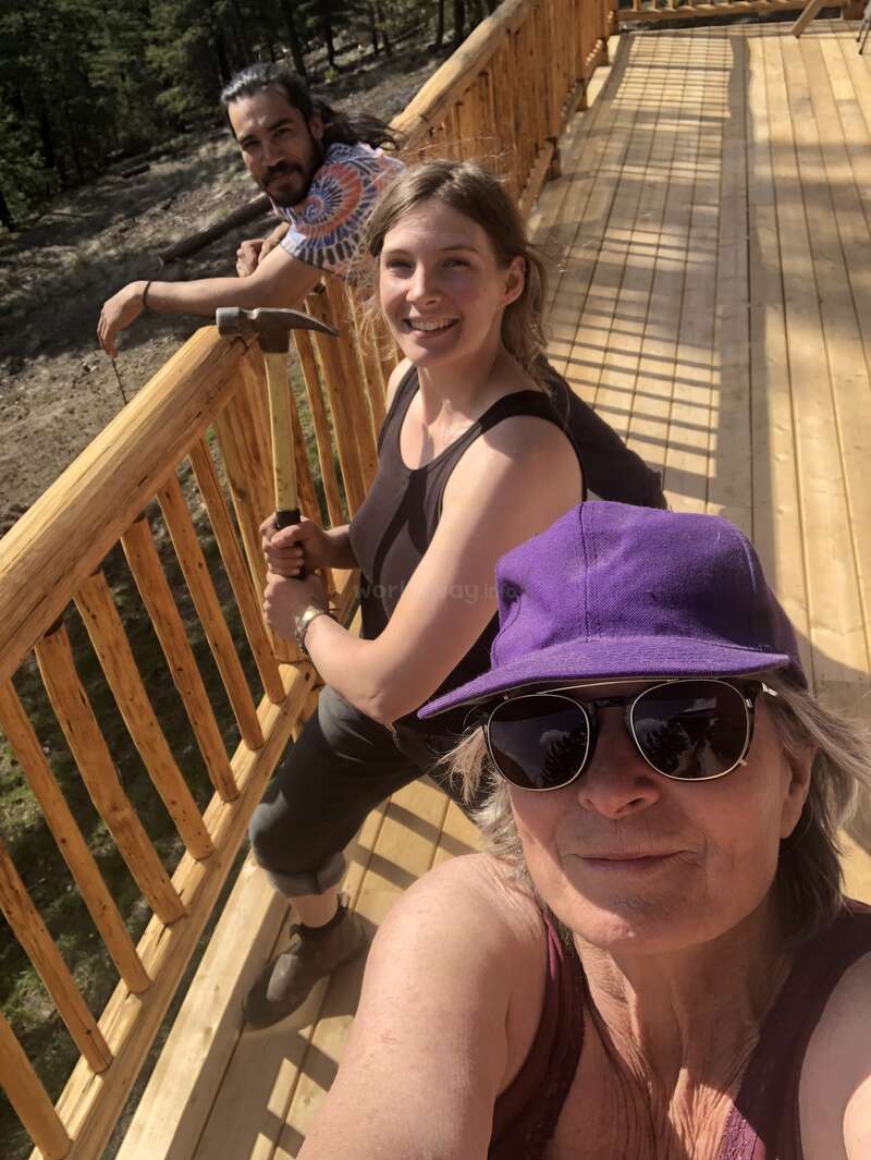 A imagem mostra três pessoas em um deck de madeira com uma grade, posando para uma selfie em uma área arborizada, com árvores e sujeira visíveis ao fundo.