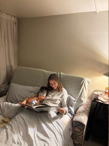 A woman and a child are cuddled up in bed, reading a large storybook together. The cozy room is softly lit by a lamp on a nightstand.