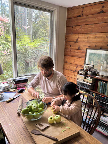 Um homem e uma jovem descascam maçãs verdes juntos em uma mesa de madeira, cercados por livros, grandes janelas e a natureza do lado de fora, criando uma atmosfera aconchegante e caseira.