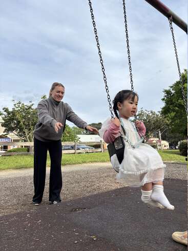 Une femme sourit et pousse une jeune fille sur une balançoire dans une aire de jeux. La jeune fille a l'air heureuse, elle porte une robe blanche. C'est une scène joyeuse.