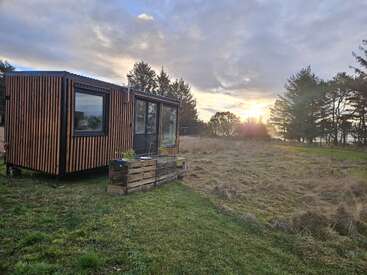 Una casita moderna con listones de madera se asienta sobre la hierba verde, rodeada de árboles, bajo un cielo espectacular al atardecer. La escena es tranquila y remota.