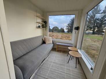 Esta acogedora habitación cuenta con un moderno sofá gris, dos pequeñas mesas redondas, un banco, grandes ventanas con vistas panorámicas al exterior y estantes de pared minimalistas con decoración.