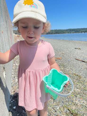 Une jeune enfant en robe rose et chapeau de soleil se tient sur une plage rocheuse, tenant un petit panier vert, avec un ciel bleu et de l'eau en arrière-plan.