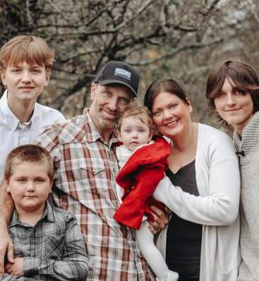 L'image représente une famille de six personnes posant pour une photo, le père portant une chemise à carreaux et une casquette de base-ball, et la mère tenant un bébé dans une veste rouge.