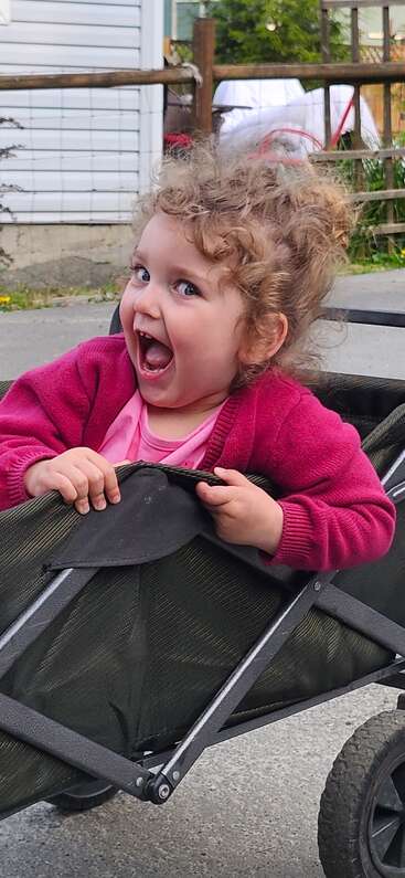 Une petite fille joyeuse aux cheveux bouclés, vêtue d'un pull rose, rit de bon cœur, assise dans une petite remorque verte à l'extérieur, par une journée ensoleillée.