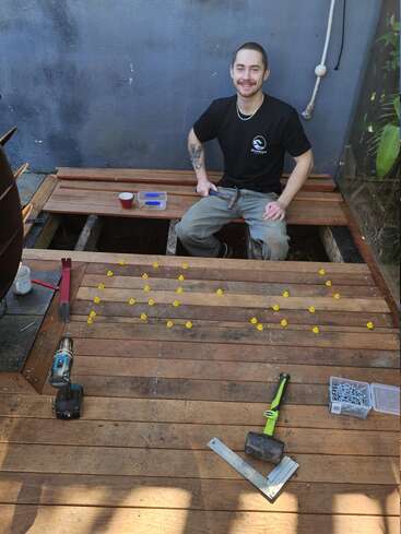 A smiling man works on installing wooden decking. Tools, screws, and yellow spacers are arranged around him. The deck is partially built, with exposed beams.