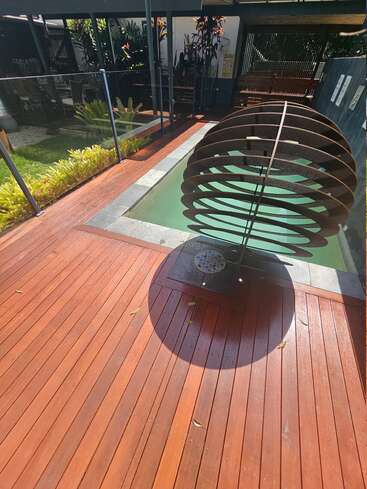 A modern outdoor area features a small pool, wooden deck, glass railing, lush greenery, and a large circular metal sculpture casting an interesting shadow.