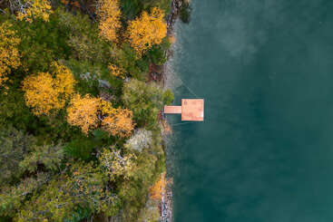 Dieses Luftbild zeigt einen Holzsteg, der sich von einer lebhaften Uferlinie mit grün-gelben Herbstbäumen in das ruhige, blaugrüne Wasser erstreckt und ein Gefühl von Ruhe und Natur vermittelt.