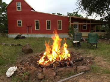 Un feu de camp vif brûle devant une maison rouge dotée de plusieurs fenêtres. Des chaises de camping vertes entourent le feu, évoquant une réunion conviviale en plein air.