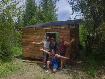 Four people celebrate in front of a small wooden cabin they built together, surrounded by lush green trees under a partly cloudy sky, smiling joyfully.