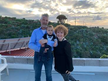 A man, woman, and baby stand together on a rooftop at sunset, surrounded by green hills and cloudy skies, exuding happiness and warmth.