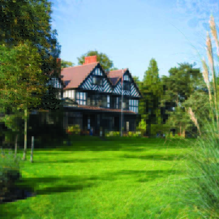 The image depicts a large, two-story house with a brown roof and white walls, surrounded by lush greenery and a well-manicured lawn on a sunny day.