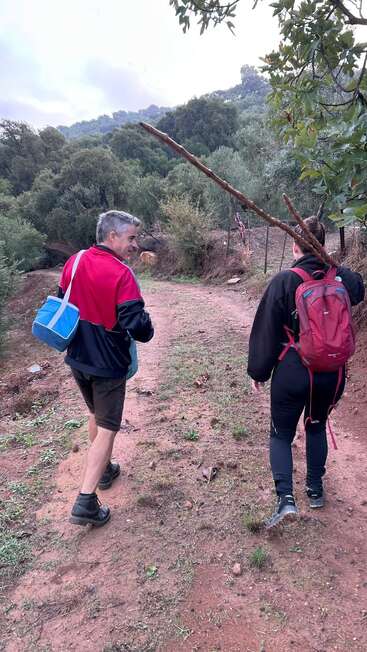 Duas pessoas caminham por uma trilha de terra em uma floresta. Uma delas carrega um bastão e uma mochila vermelha; a outra tem uma bolsa azul e usa botas.