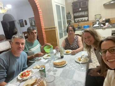 Cinco pessoas estão sentadas ao redor de uma mesa de jantar, compartilhando uma refeição em uma cozinha aconchegante. Todos parecem felizes, apreciando a comida e a companhia uns dos outros.