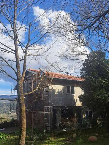 A two-story house under construction or renovation, surrounded by scaffolding and trees. Bright blue sky with white clouds, green grass, and distant hills are visible.