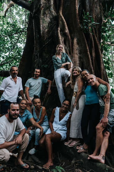 A group of eleven people pose happily in front of a massive tree with sprawling roots, surrounded by lush greenery. The atmosphere feels relaxed and friendly.